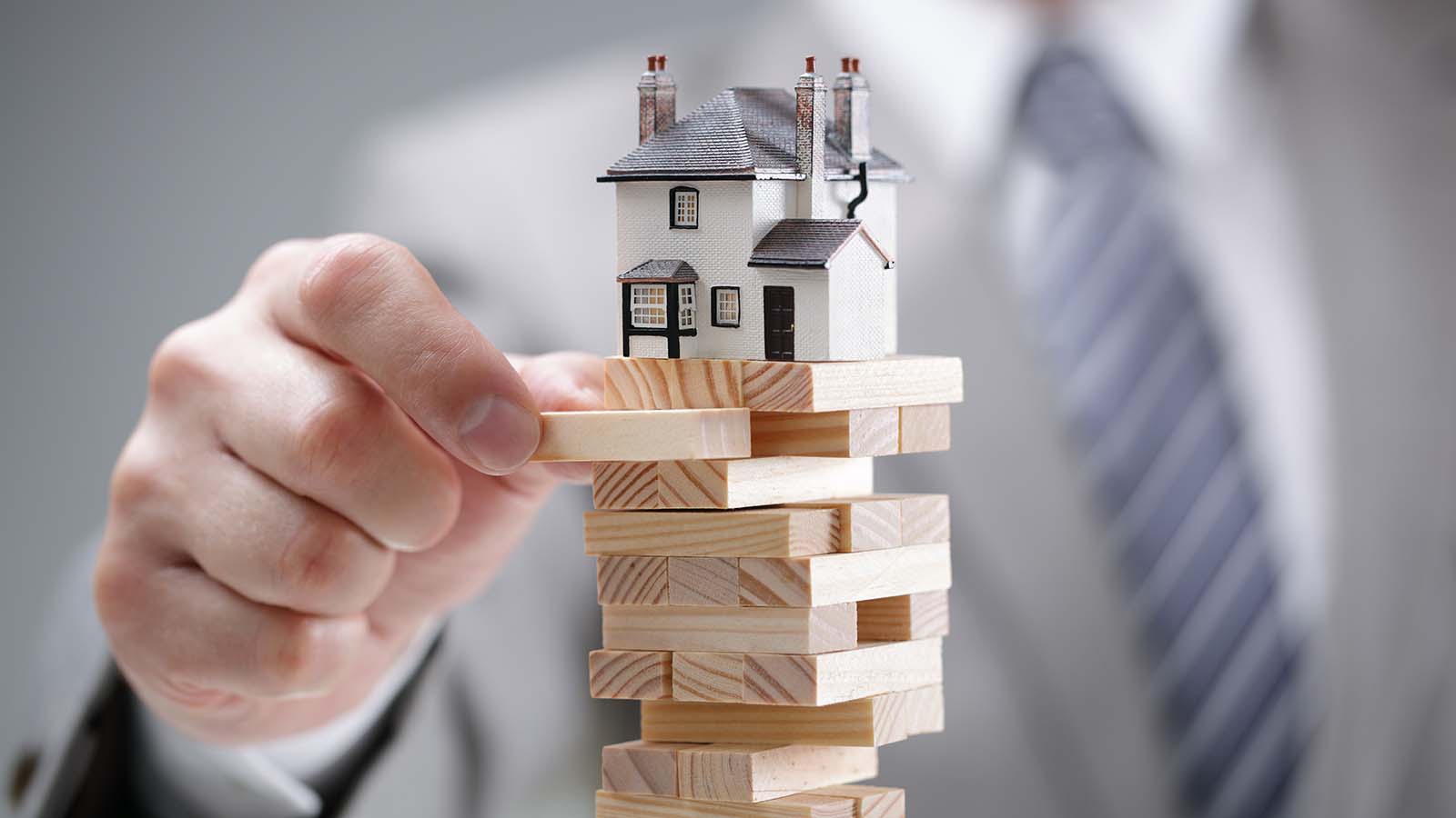A close-up shot of a hand pulling out a Jenga block with a model house sitting on top of the tower.