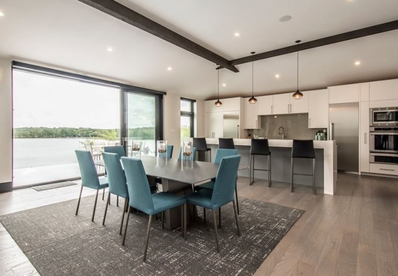 A dining room and kitchen combo.  There is a large dining table in front of a kitchen island that has four barstool seats.  There is a large window overlooking a lake at the end of the table on the left side.